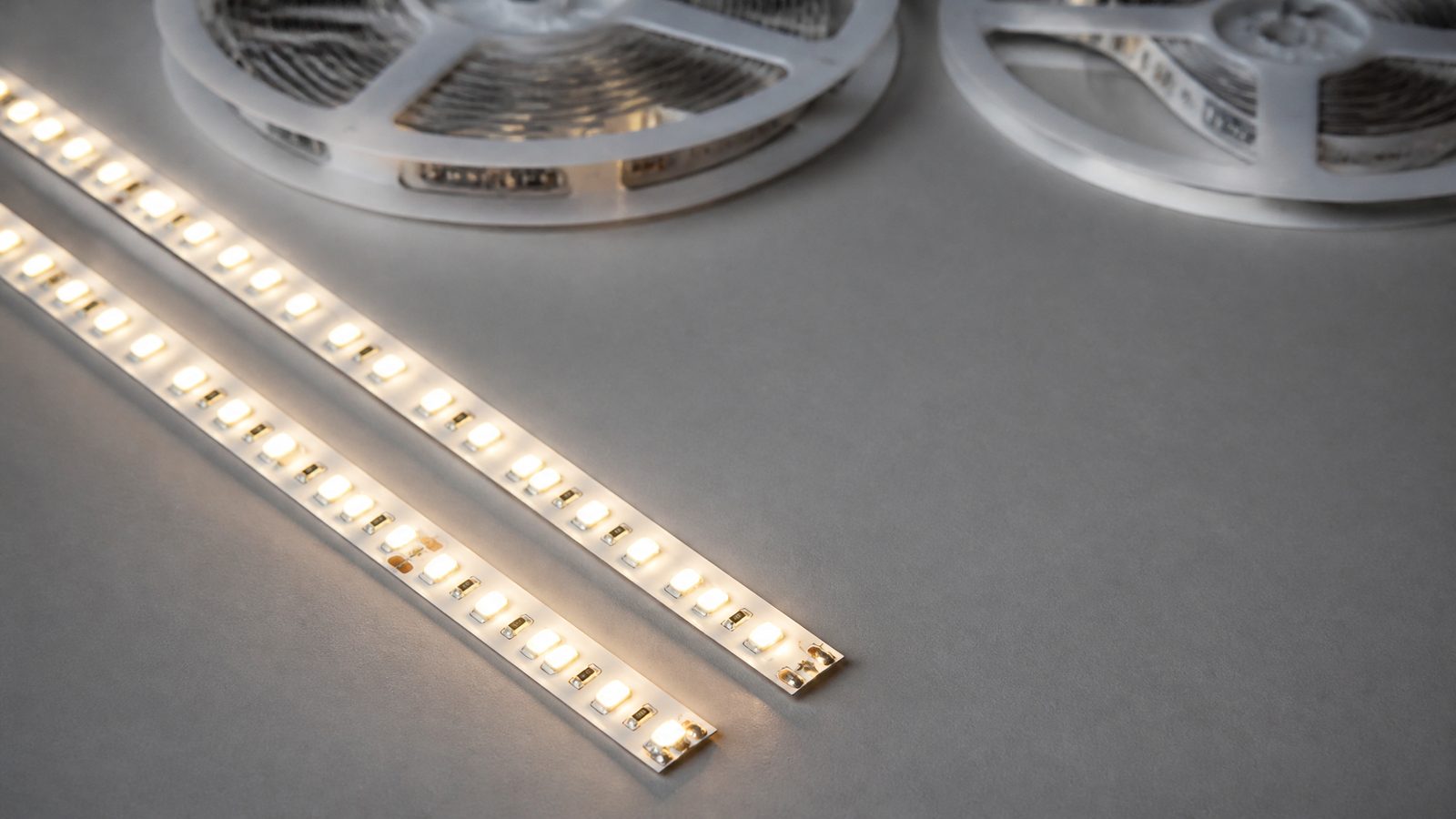 Two lit warm-white LED strips side by side on a gray surface, with unlit reels behind them.