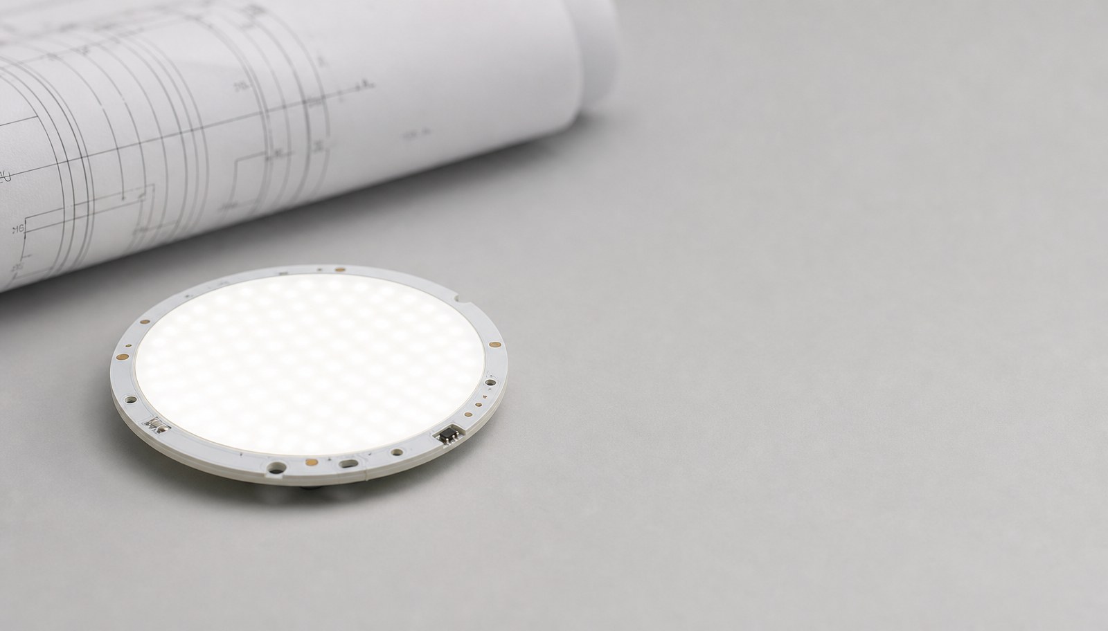 A small round illuminated LED light board module beside a rolled technical drawing sheet.